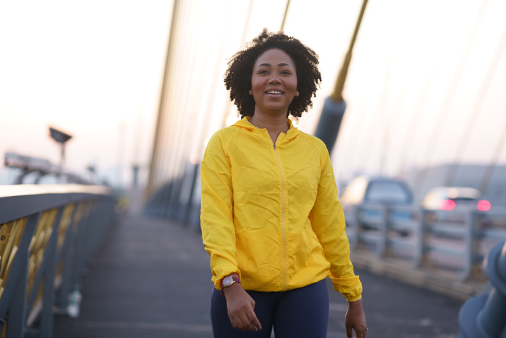A young woman in a yellow jacket walking on a pedestrian bridge, with cars in the background.