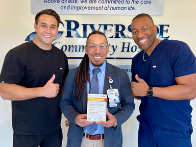 Smiling hospital staff pose in front of hospital sign while holding the Environmental Excellence Award and give thumbs up.