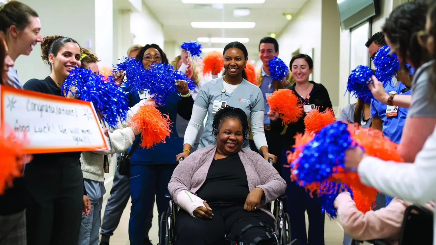 Patient in a wheelchair being pushed down a hallway surrounded by nurses holding pompoms and congratulation signs.