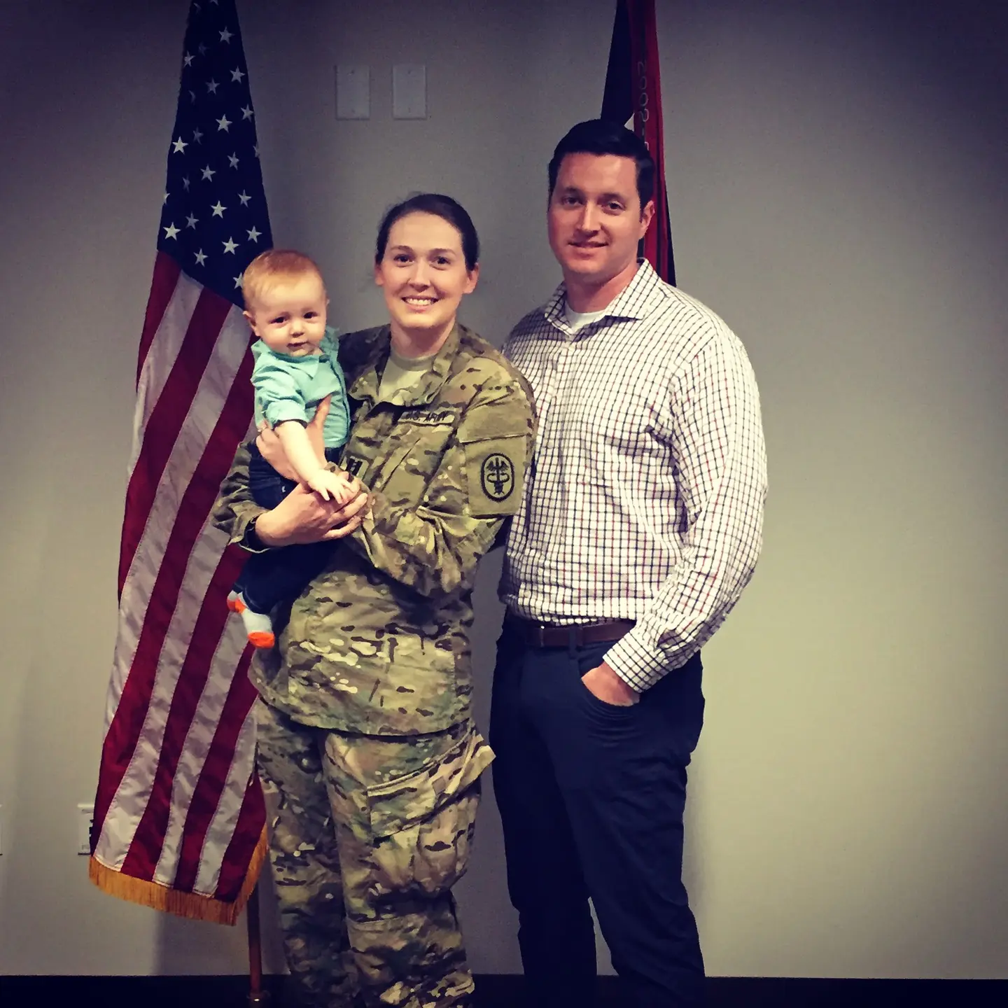 Taylor Maynard holds her son and stands next to her husband in front of the American flag.