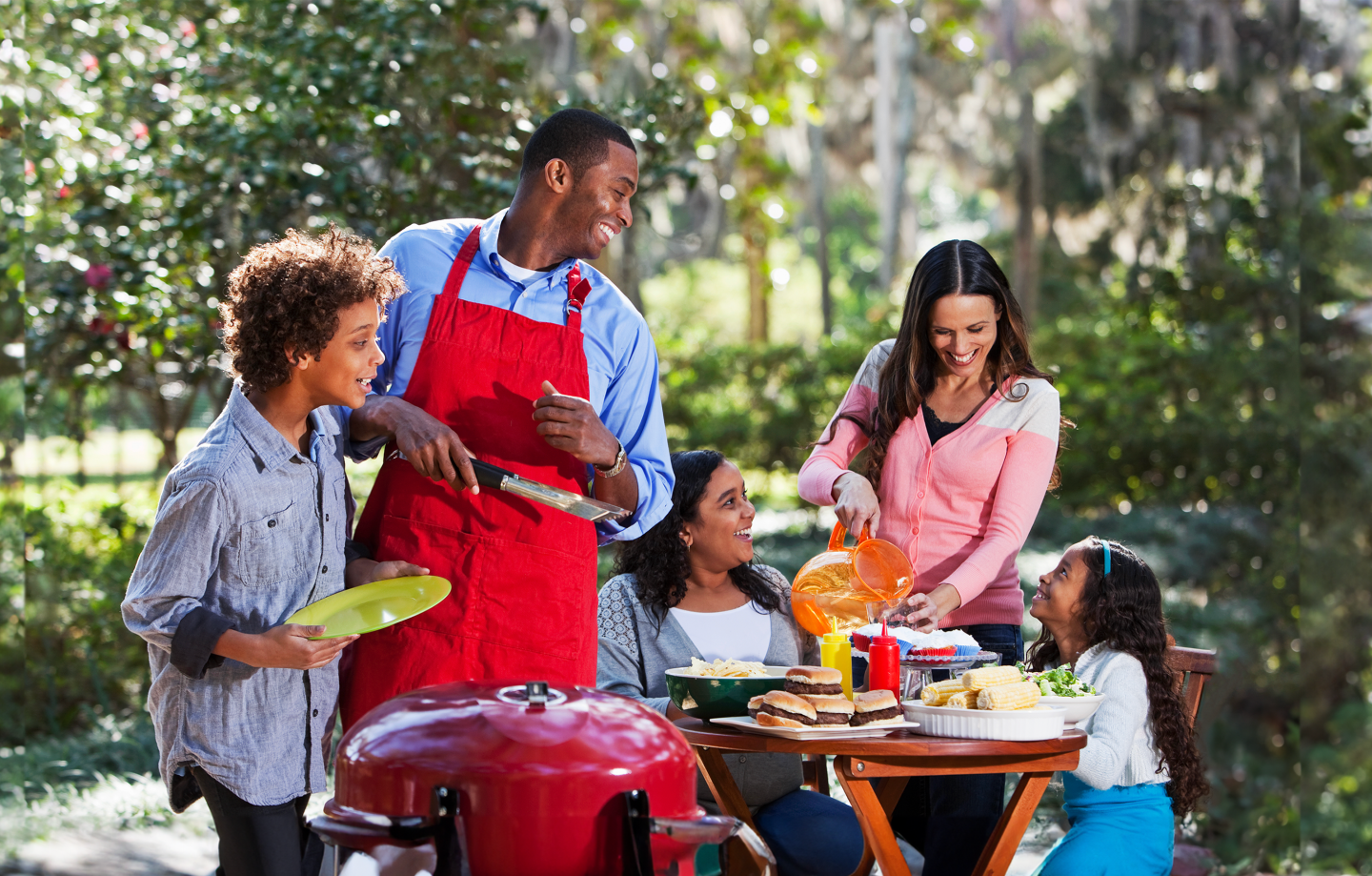 A smiling family of 5, having a barbeque outdoors, gathered around a patio table, preparing to eat burgers.