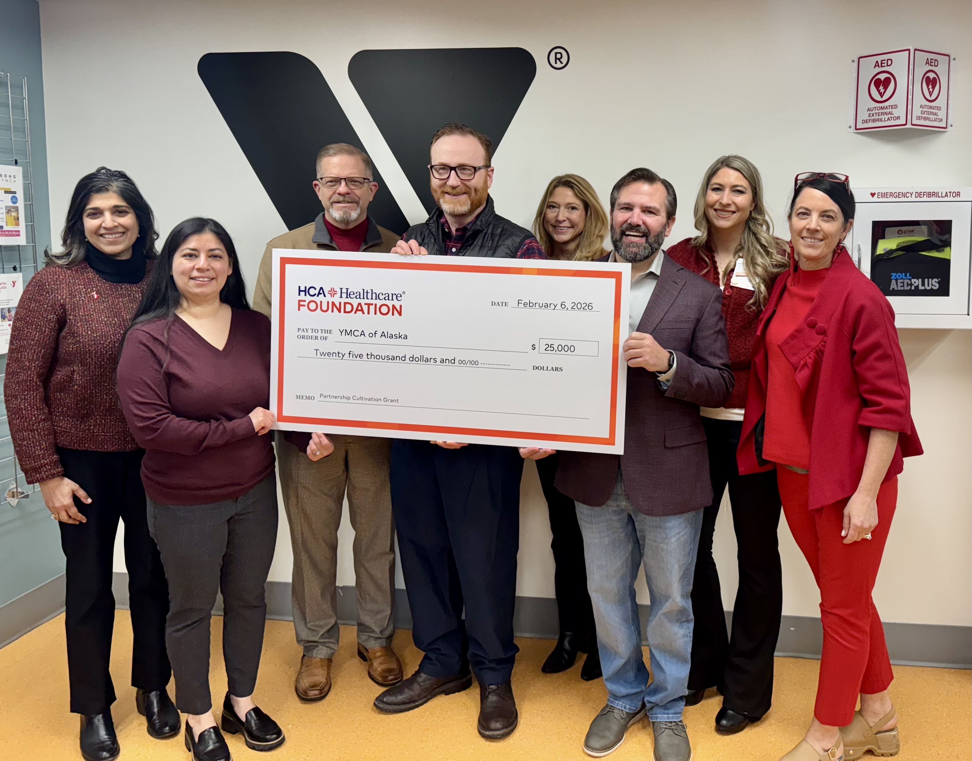 Members of the administration team at Alaska Regional Hospital and representatives of the YMCA of Alaska all pose together in a group, smiling, at the Anchorage Y location. Members of the group are wearing red and holding up a large check -- a $25,000 grant from the HCA Healthcare Foundation made out to the YMCA of Alaska.