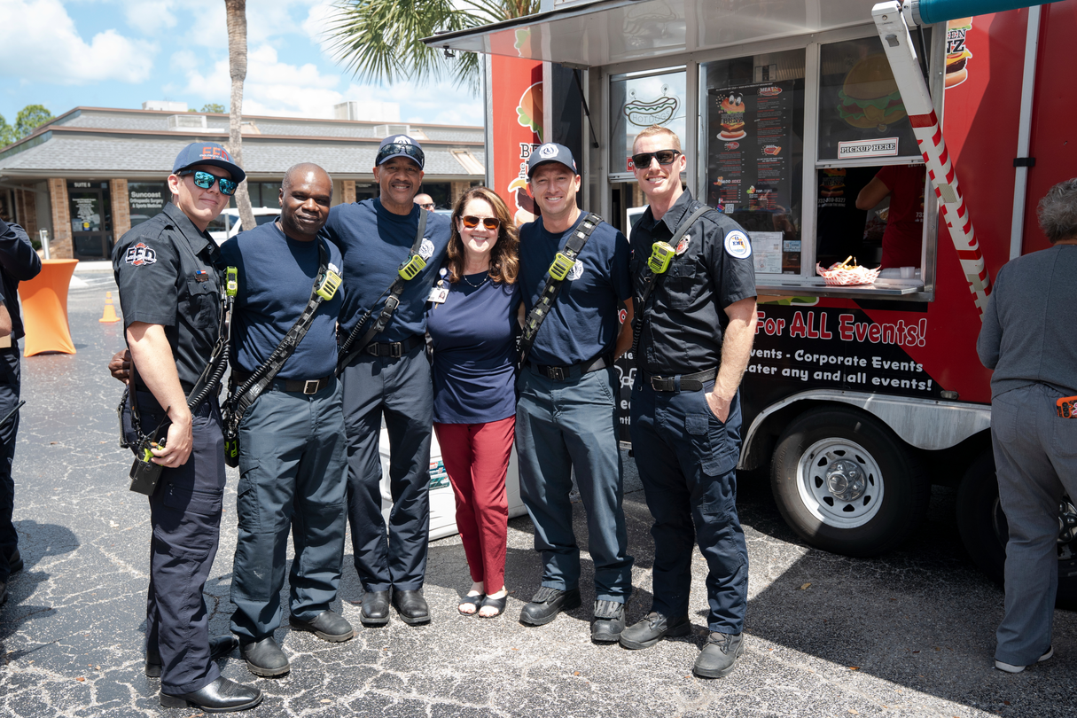 Five first responders and a hospital team member pose together outside a food truck during an EMS Week celebration at HCA Florida Englewood Hospital. The group stands in front of the truck on a sunny day, recognizing the partnership between emergency responders and healthcare teams.