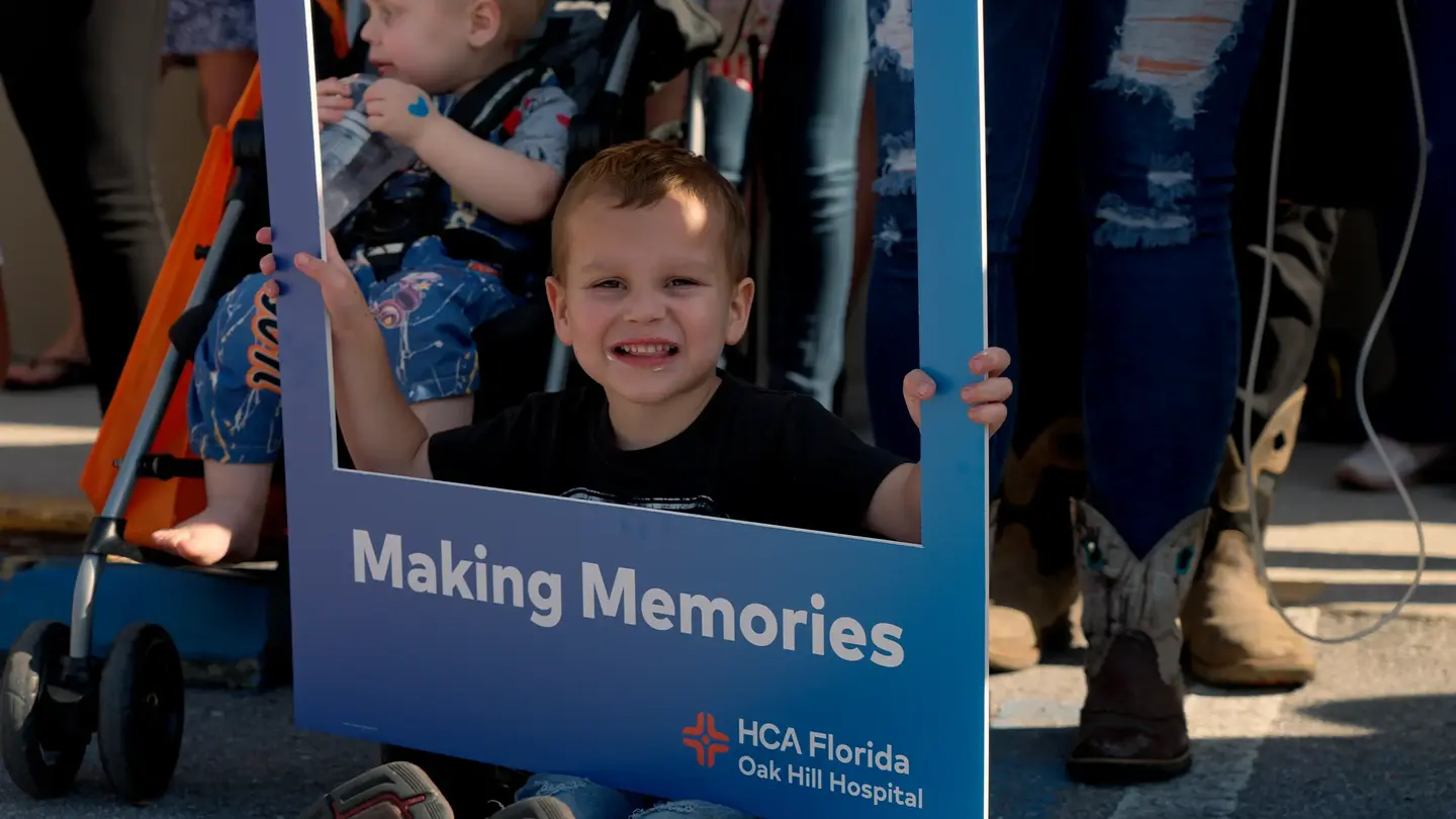 Child smiling and looking thru an HCA Florida Oak Hill Hospital cutout board that says "Making Memories"