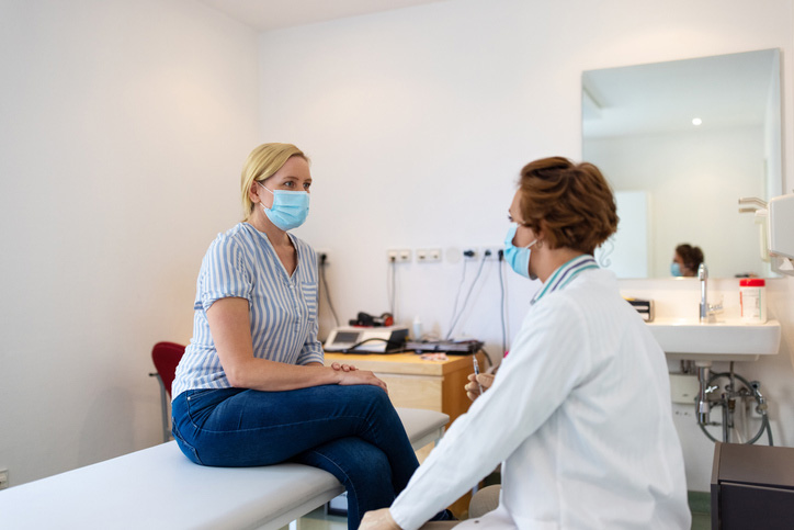 A doctor talks with a female patient in an exam room, while both wear face masks.