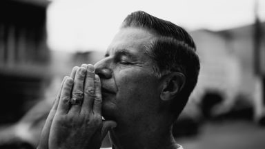 A man holds his folded hands up to his mouth, with his eyes closed.