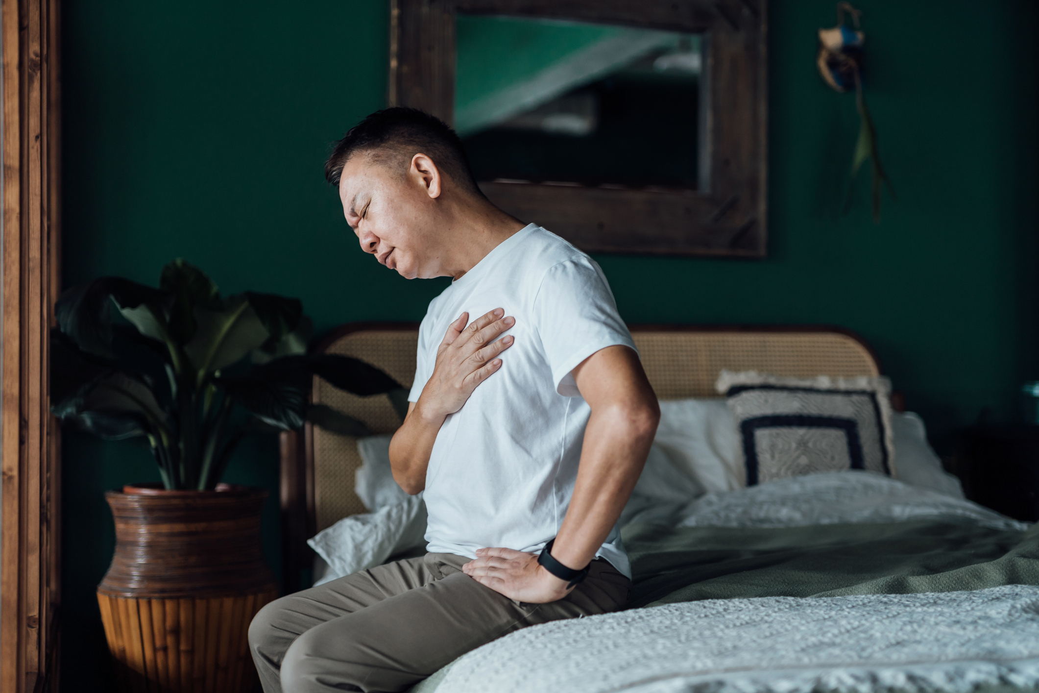 A man sits on a bed with his eyes closed, holding his chest in discomfort.