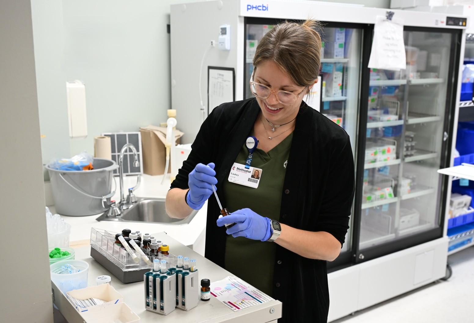 A Methodist Hospital Texsan colleague wearing gloves uses a pipette to transfer liquid from a small vial while working in a clinical laboratory. Refrigerated storage units, testing supplies, and lab equipment are visible in the background.