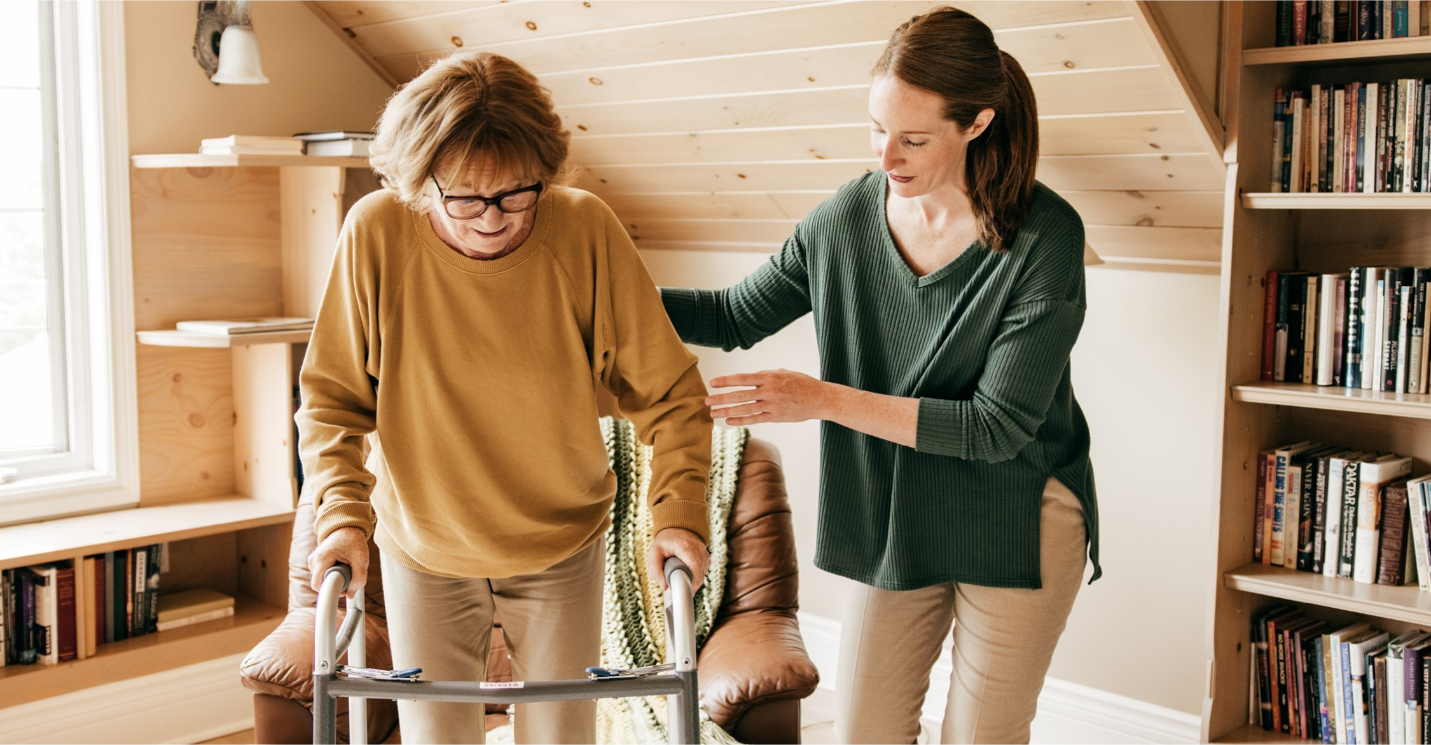 Younger woman helping older woman stand from chair with walker.