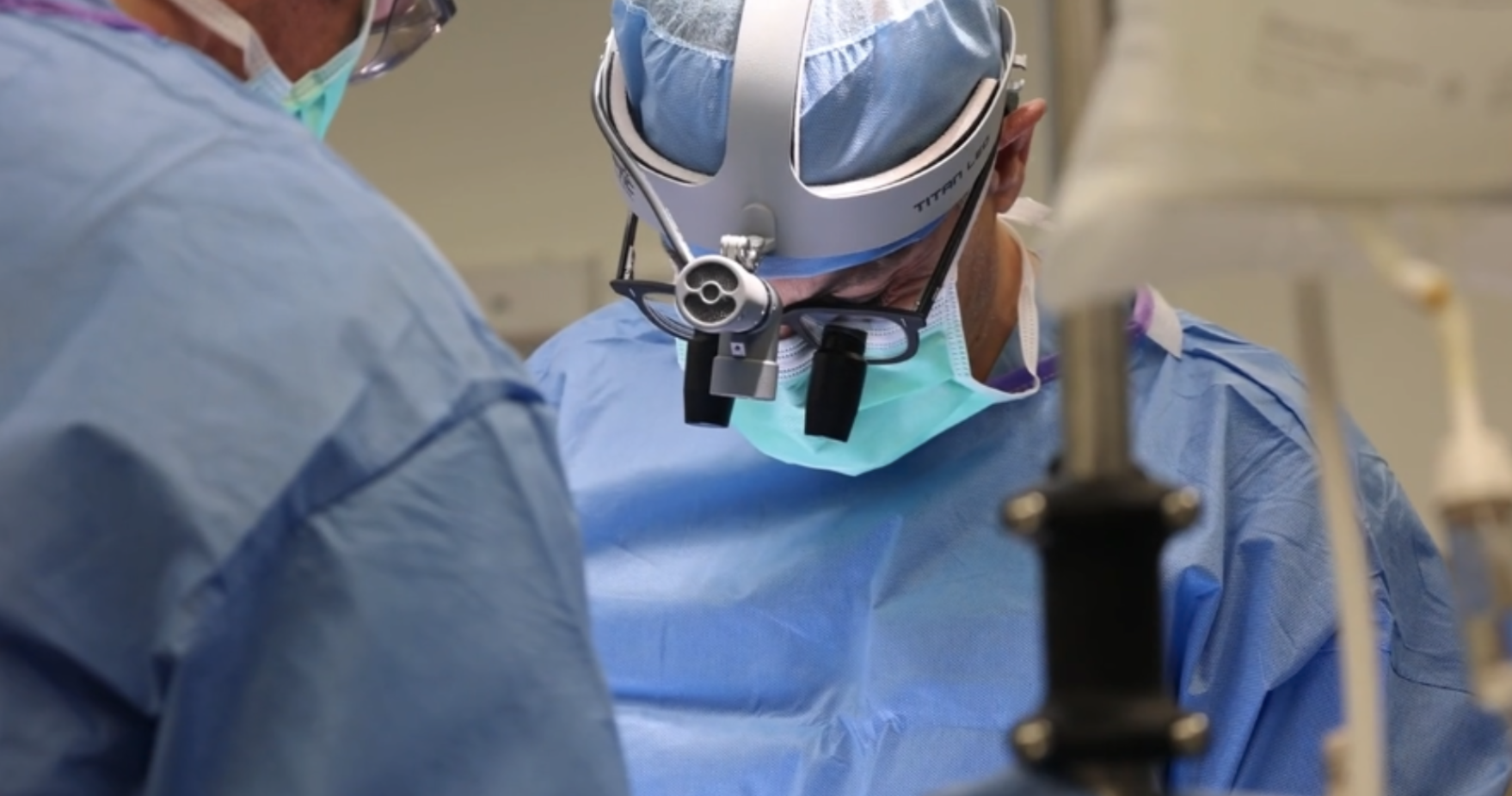 Two doctors perform surgery, while wearing blue surgical scrubs and head lamps.
