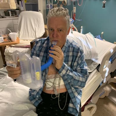 Justin Phillips on a hospital bed holding a tube to his mouth.
