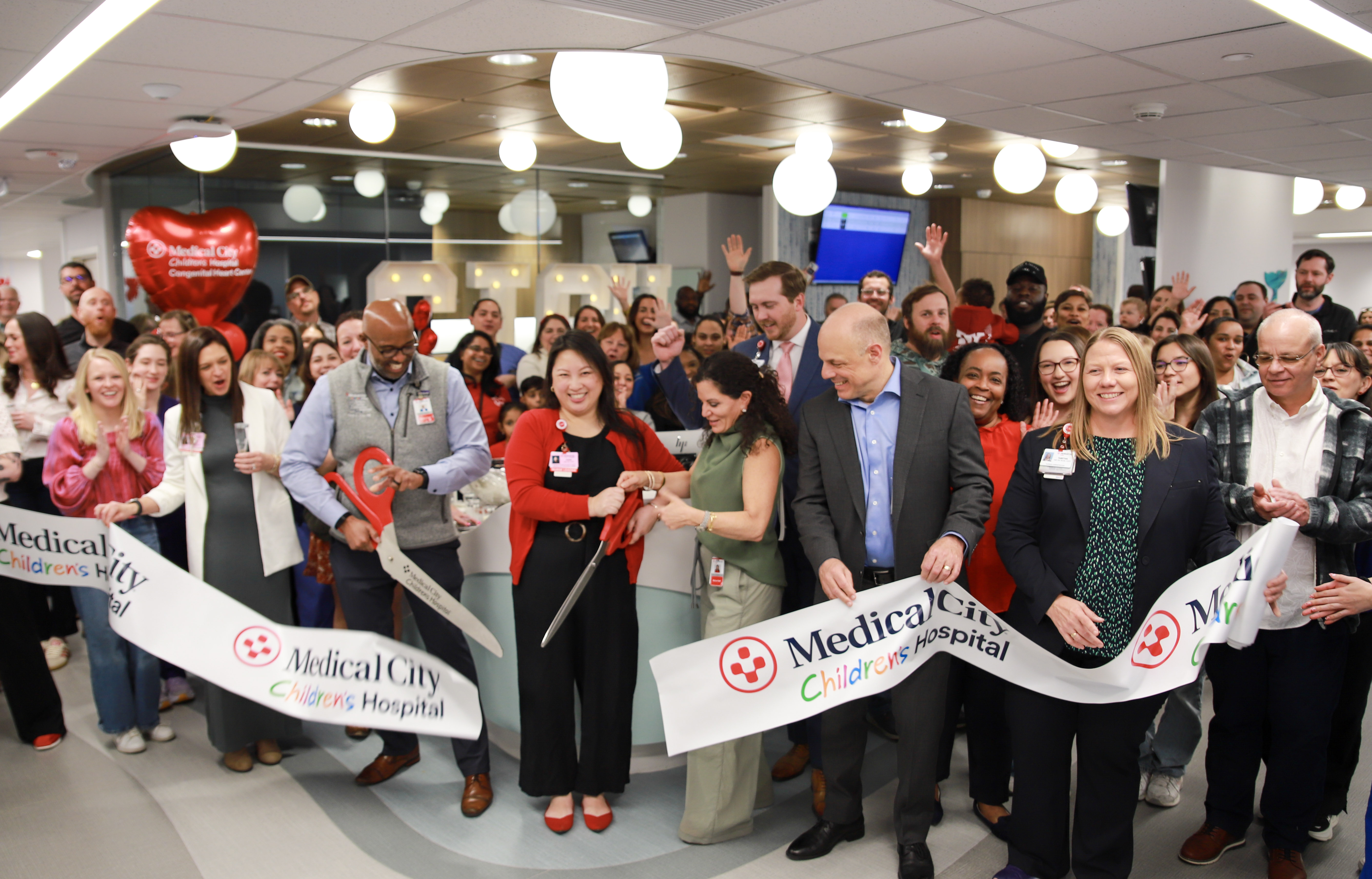 A large group of hospital leaders, staff and patients gather indoors for a ribbon-cutting ceremony at Medical City Children's Hospital, smiling and cheering as several executives use oversized scissors to cut a branded ribbon celebrating the opening of the Congenital Heart Center expansion. Red heart-shaped balloons and a crowd of applauding attendees fill the background.