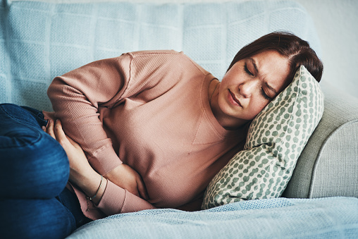 A woman laying on her side, on a couch, with her arms wrapped around her abdomen. She has an facial expression that indicates she's in pain.