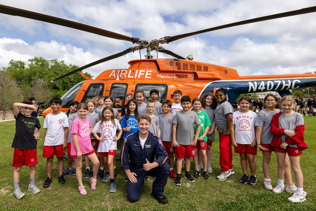 A large group of healthcare staff, officials, and community members stand on grass in front of an orange AirLife medical helicopter during a ribbon-cutting ceremony.