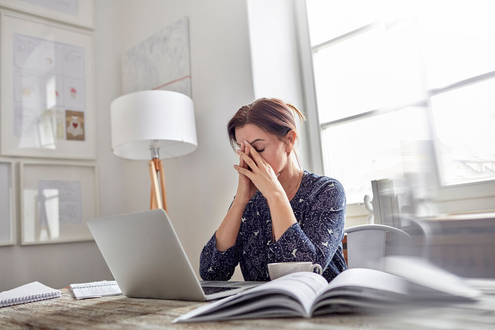 Woman sitting at a desk with a laptop, covering her face with her hands, appearing stressed or overwhelmed in a bright home office.