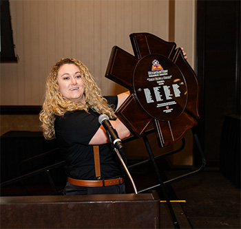A woman at a podium and microphone, proudly holds up a plaque printed with the names of the graduating students.