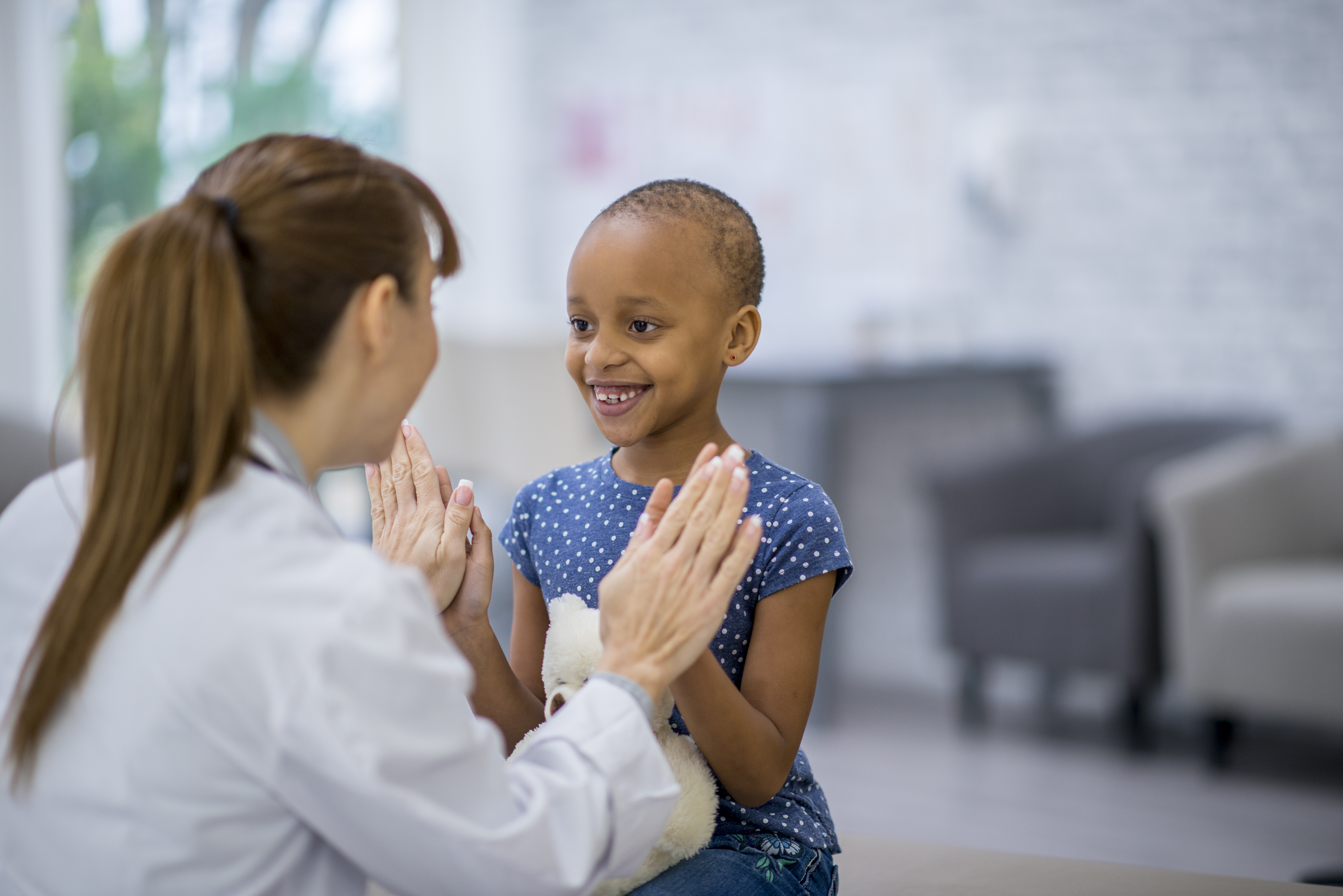A young girl with a shaved head gives a high five to a doctor, inside a hospital lobby.