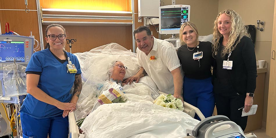 Mary, RN, Tessa, RN, and Dr. Macdonald beside patient Lori, in her hospital bed and wearing a wedding veil, with her husband Eddie standing to her right.