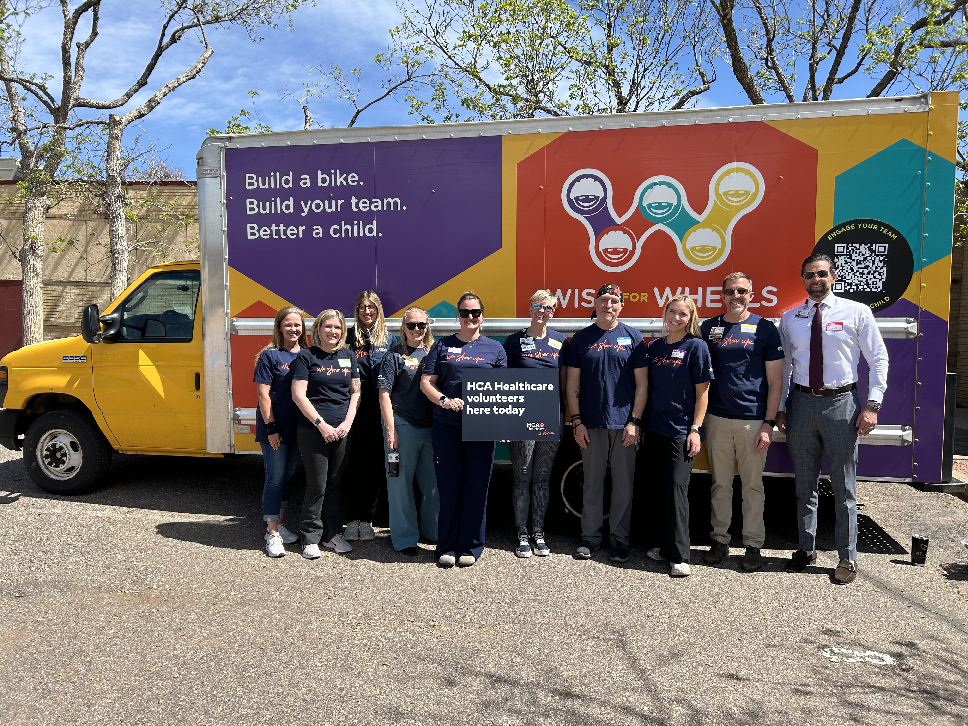 A group of volunteers standing in front of a yellow truck that says "Build a bike. Build your team. Better a child." and smiling for a picture.