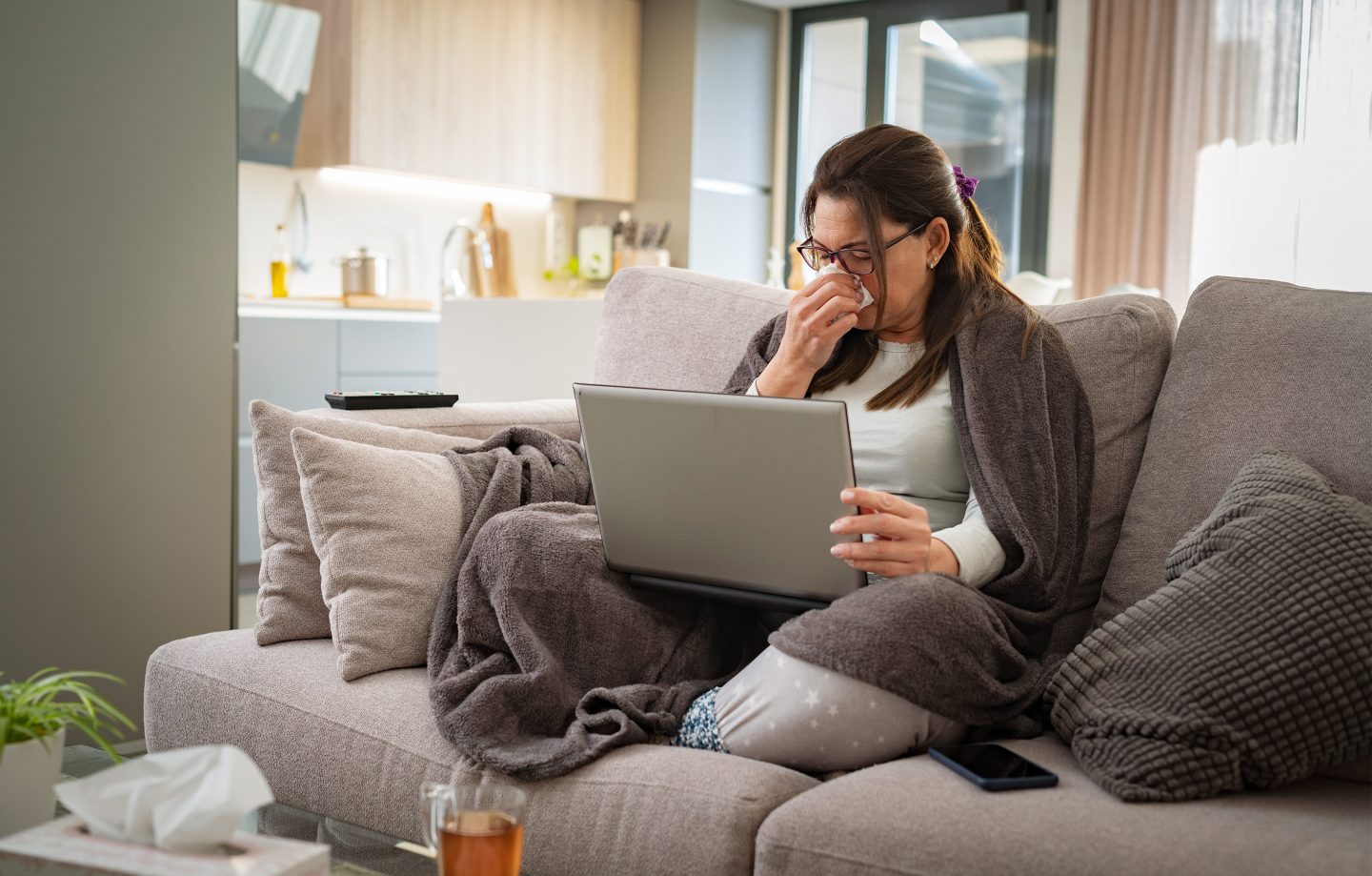 A woman on a couch with a blanket on her shoulders. She has a laptop in her lap and she's staring at it while holding a tissue to her nose.