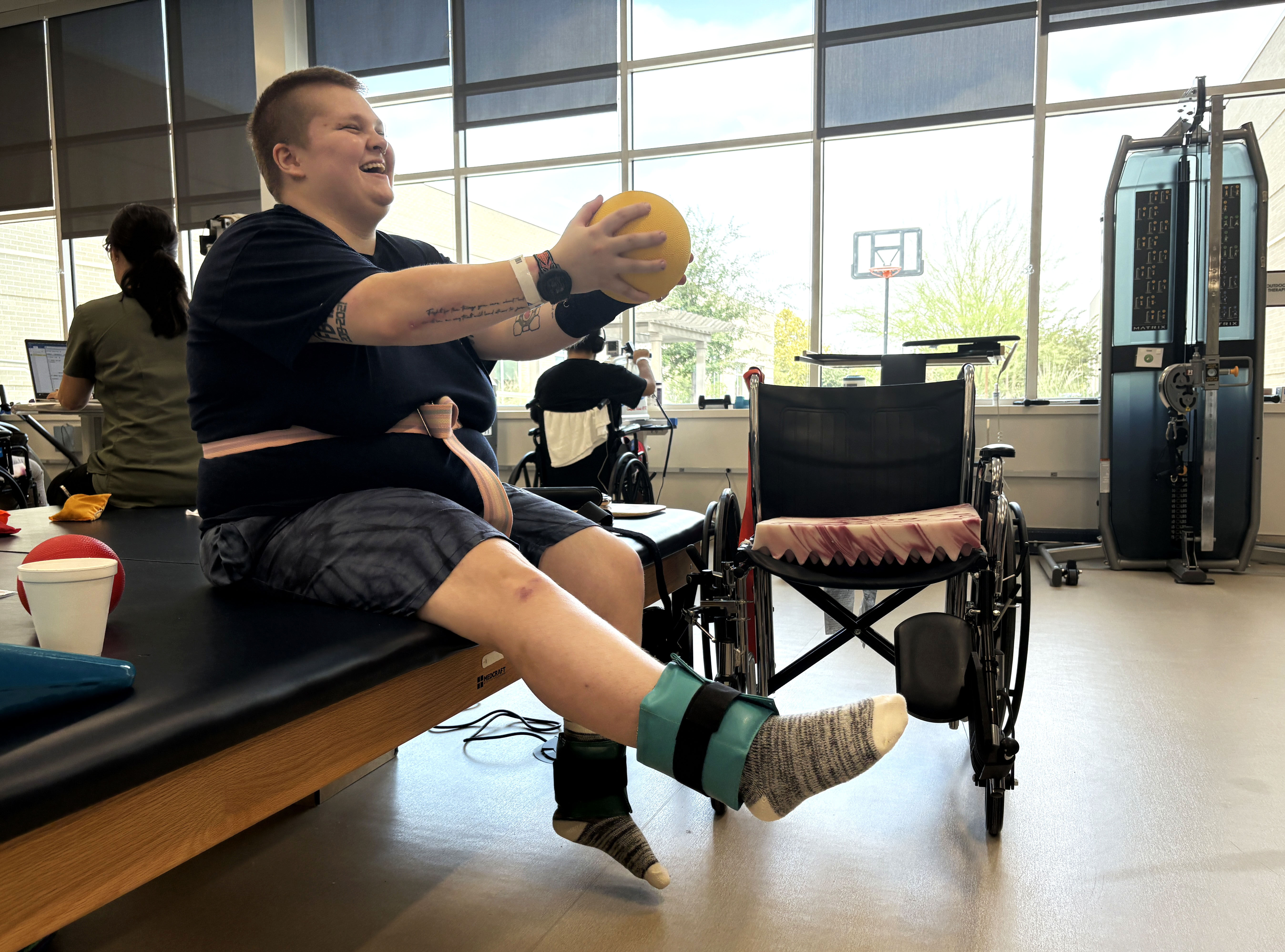 A person sits on a padded therapy table in a rehabilitation gym, holding a yellow exercise ball with both hands while extending one leg forward. The leg has a green ankle weight, and the person is wearing gray socks. A wheelchair is positioned nearby, and exercise equipment is visible in the background near large windows.