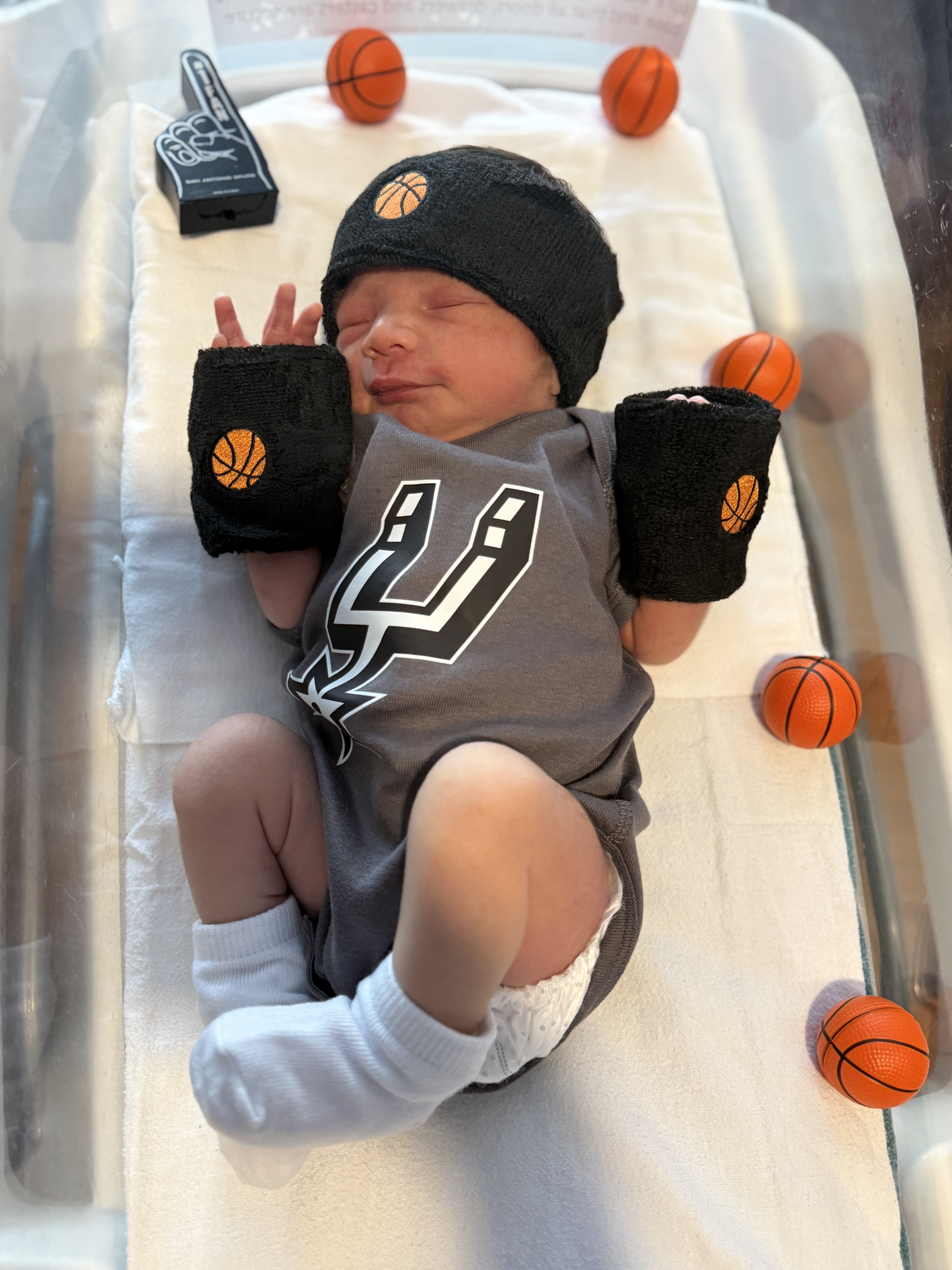 Newborn baby dressed in Spurs-themed outfit lying next to a mini basketball at a Methodist Healthcare hospital.
