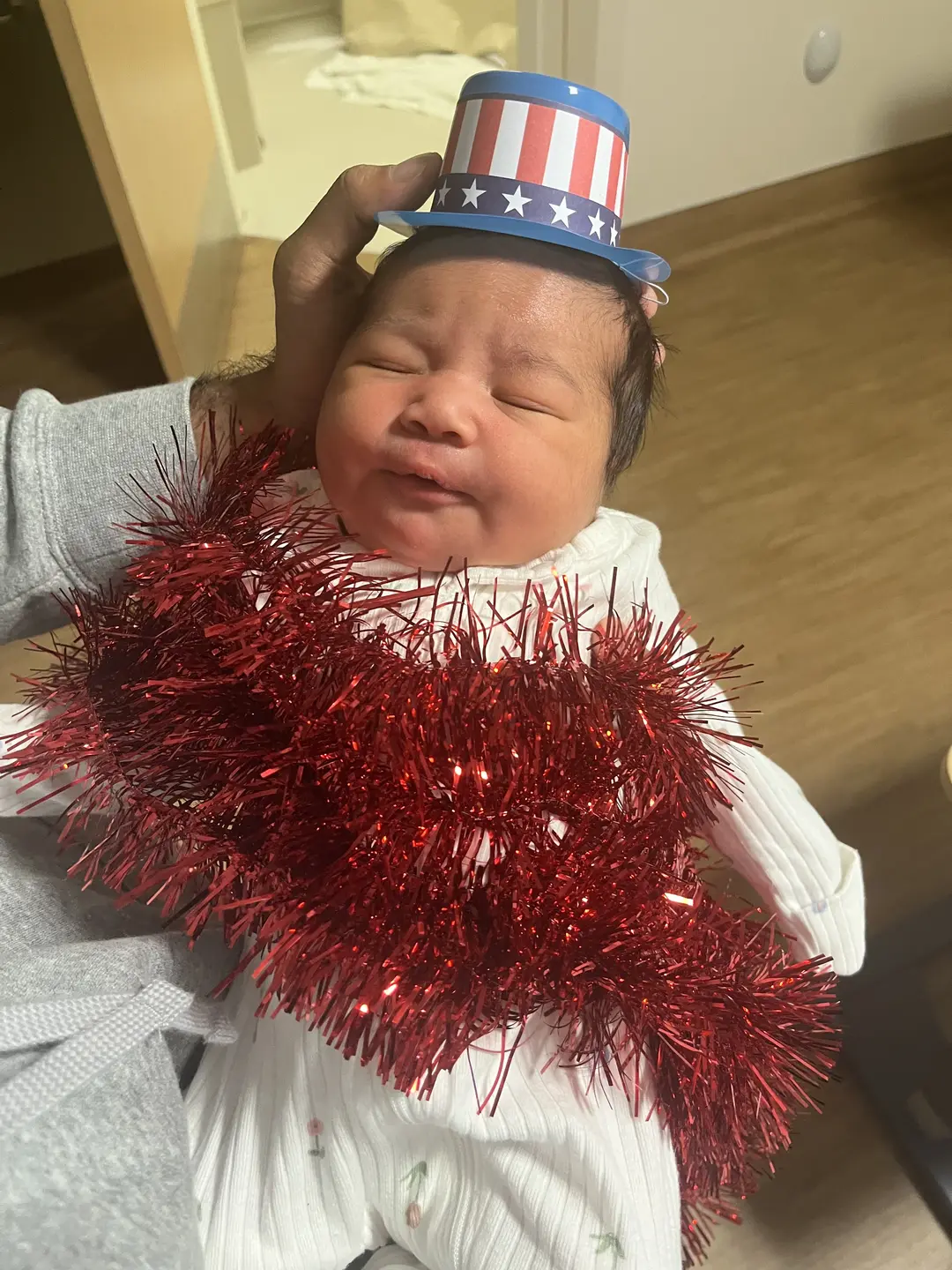 A newborn baby sleeping in a parent's arms, wearing a decorative red garland and a little patriotic top hat.