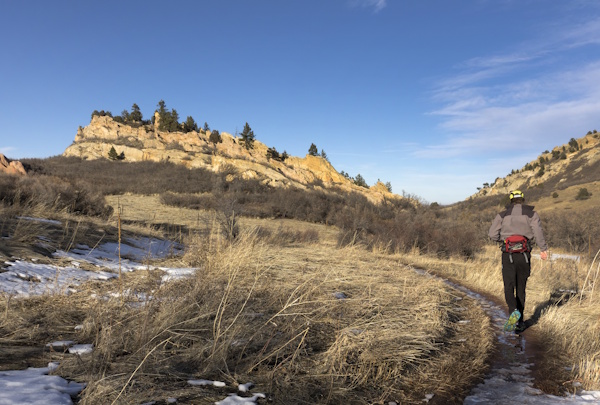Man runs trail in mountains, with melting snow around him.