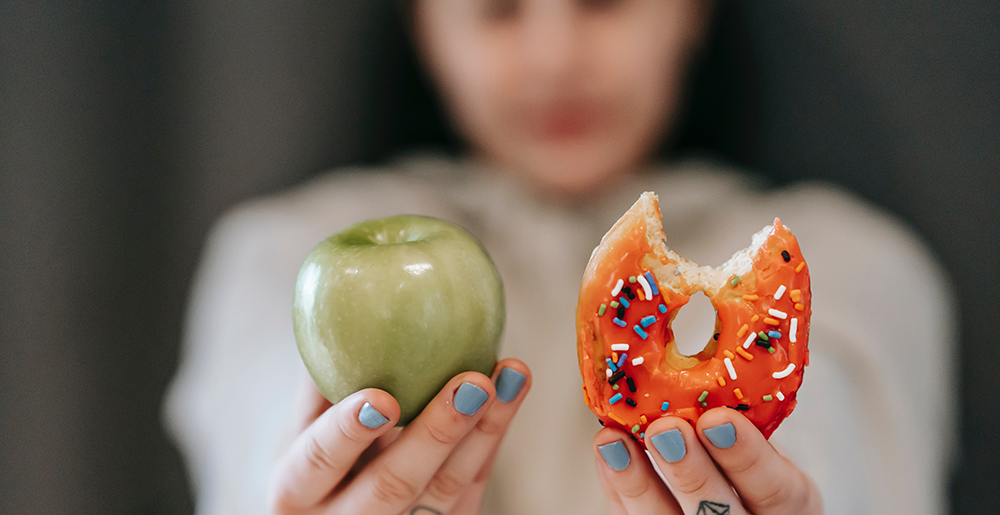 A woman wearing blue nail polish holds up a green apple and a donut next to each other.