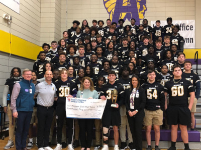 Student athletes wearing football uniforms and hospital staff and executives gather for group portrait in gym.