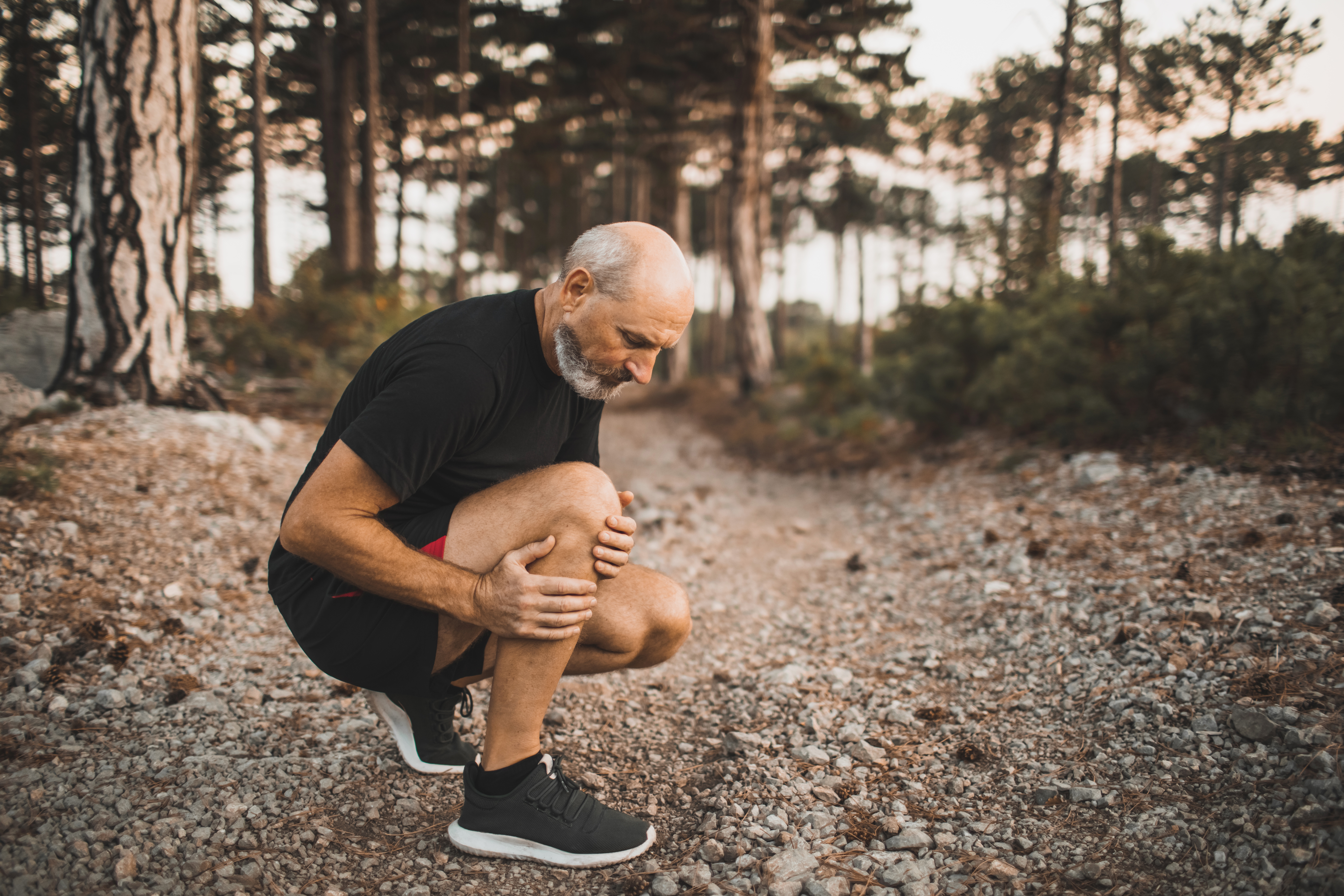 A senior man jogging through a wooded area stops to crouch and rub his right leg, indicating joint or muscle pain.