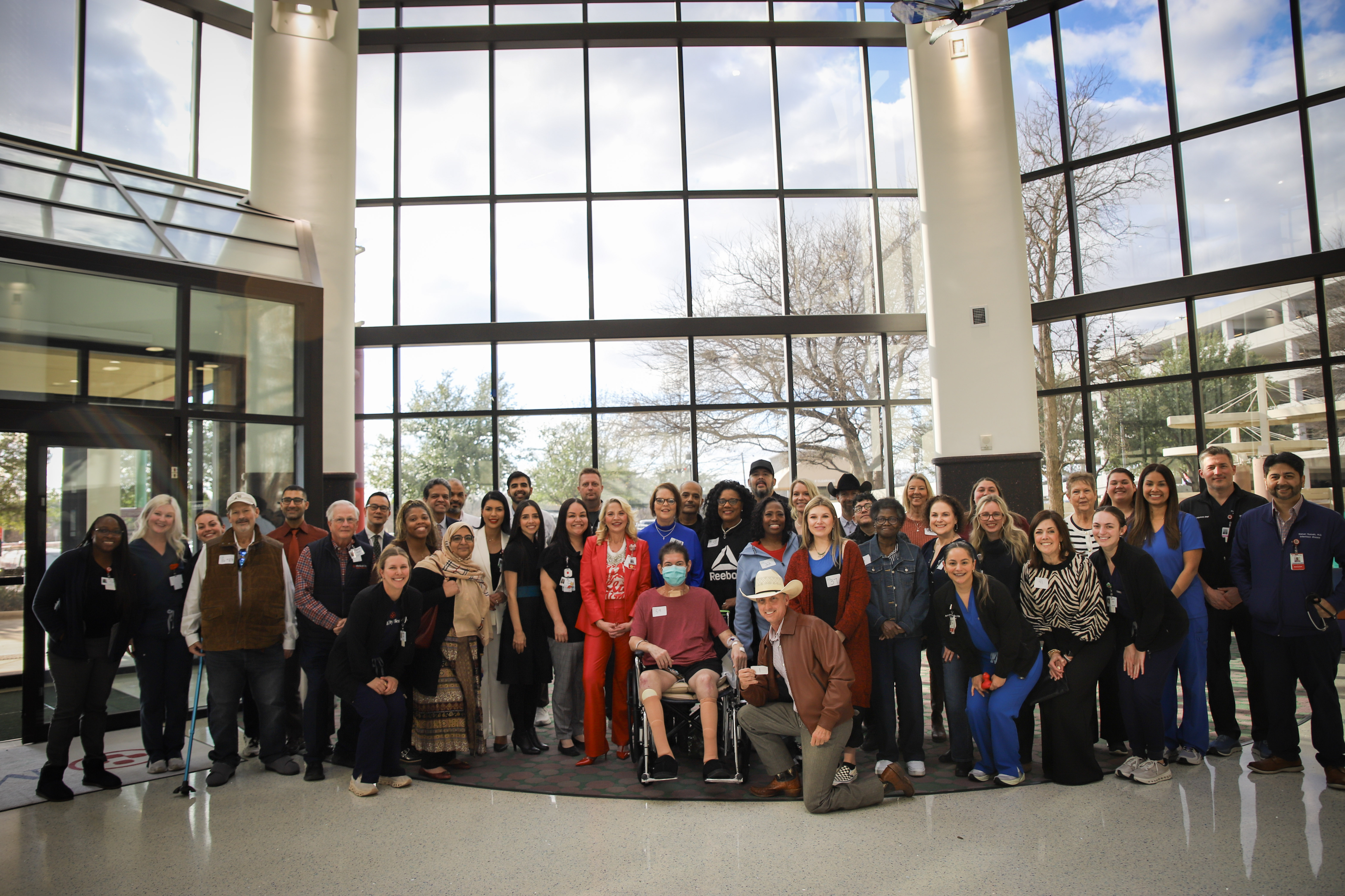 Large group of Medical City Dallas transplant patients and hospital colleagues gathered in a bright hospital lobby, celebrating the program's 100th liver transplant milestone.