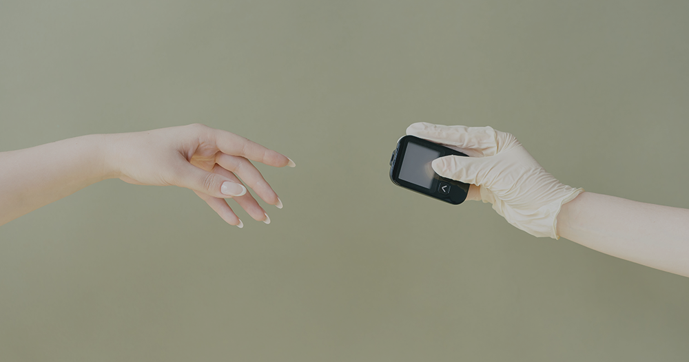 A woman's hand reaches for a diabetic testing machine being handed to her by a hand wearing a nitrile glove.