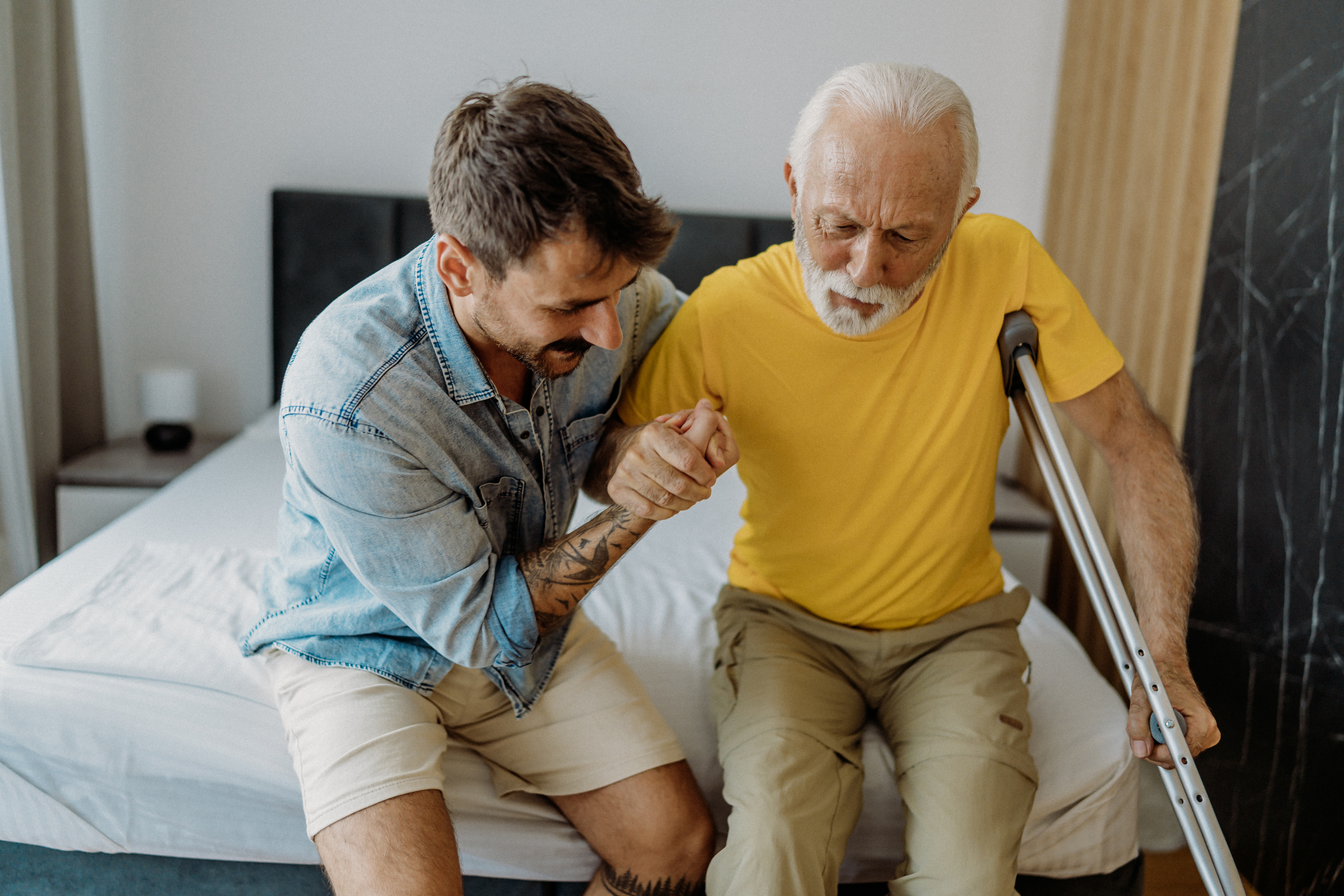 Son helping his father stand up off the bed. The father is holding a crutch under one armpit and holding onto his son's hand with the other.