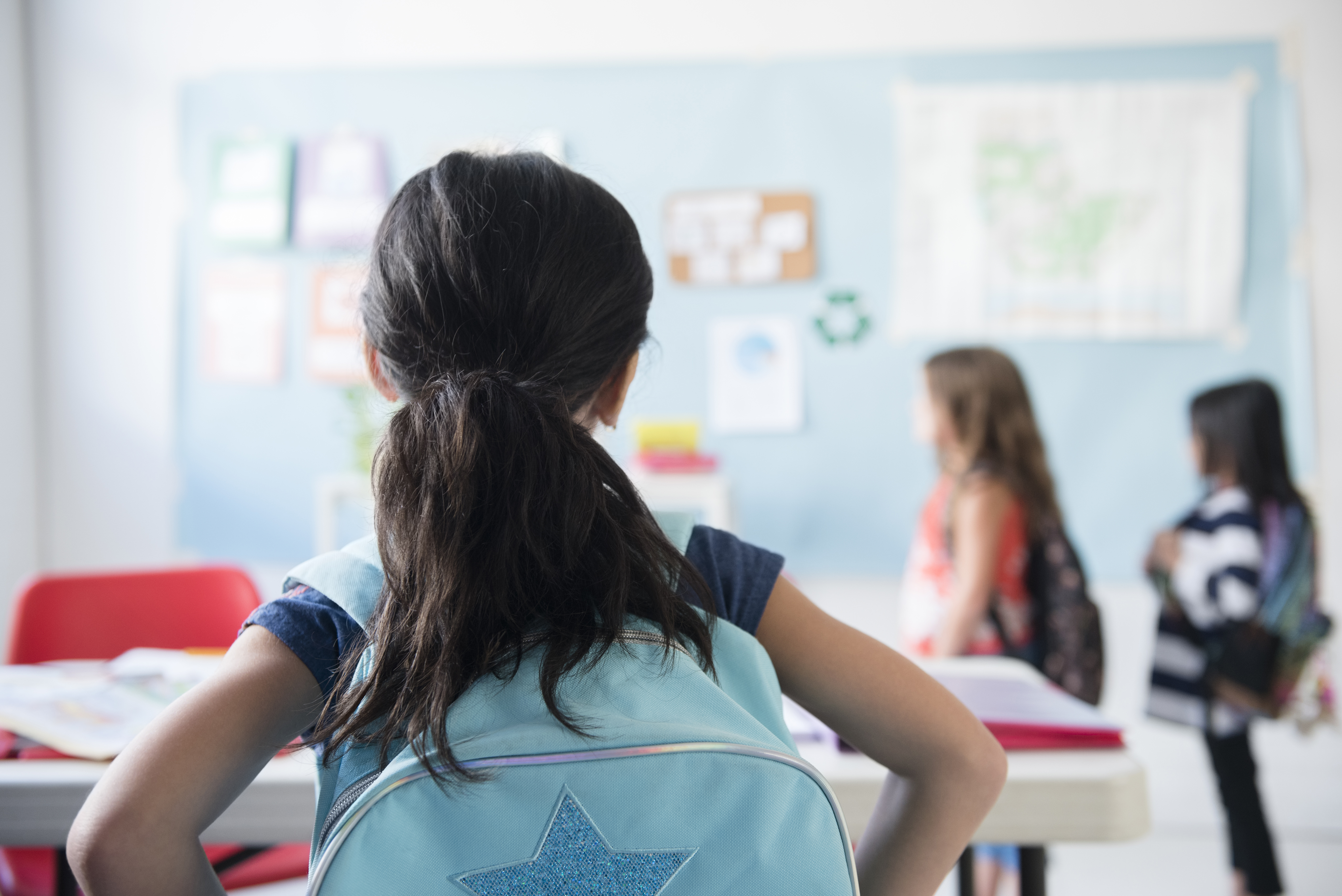 From behind, a young girl wearing a light blue backpack with a glittering star on it looks over at her classmates in a classroom.