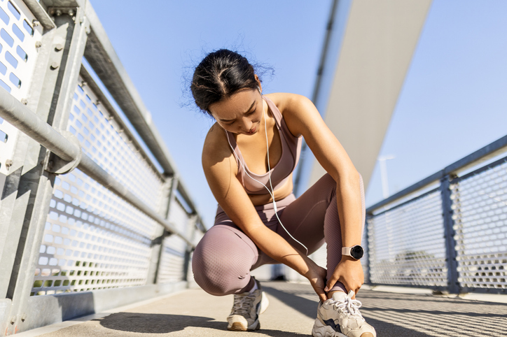 Young woman experiences a leg injury while jogging, bending down on a bridge.
