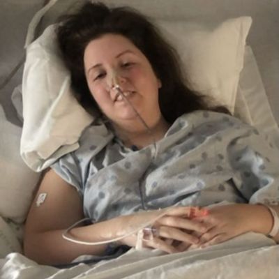 Emma Nelson smiles while sitting in a hospital bed, with several medical tubes hooked to her hand and face.
