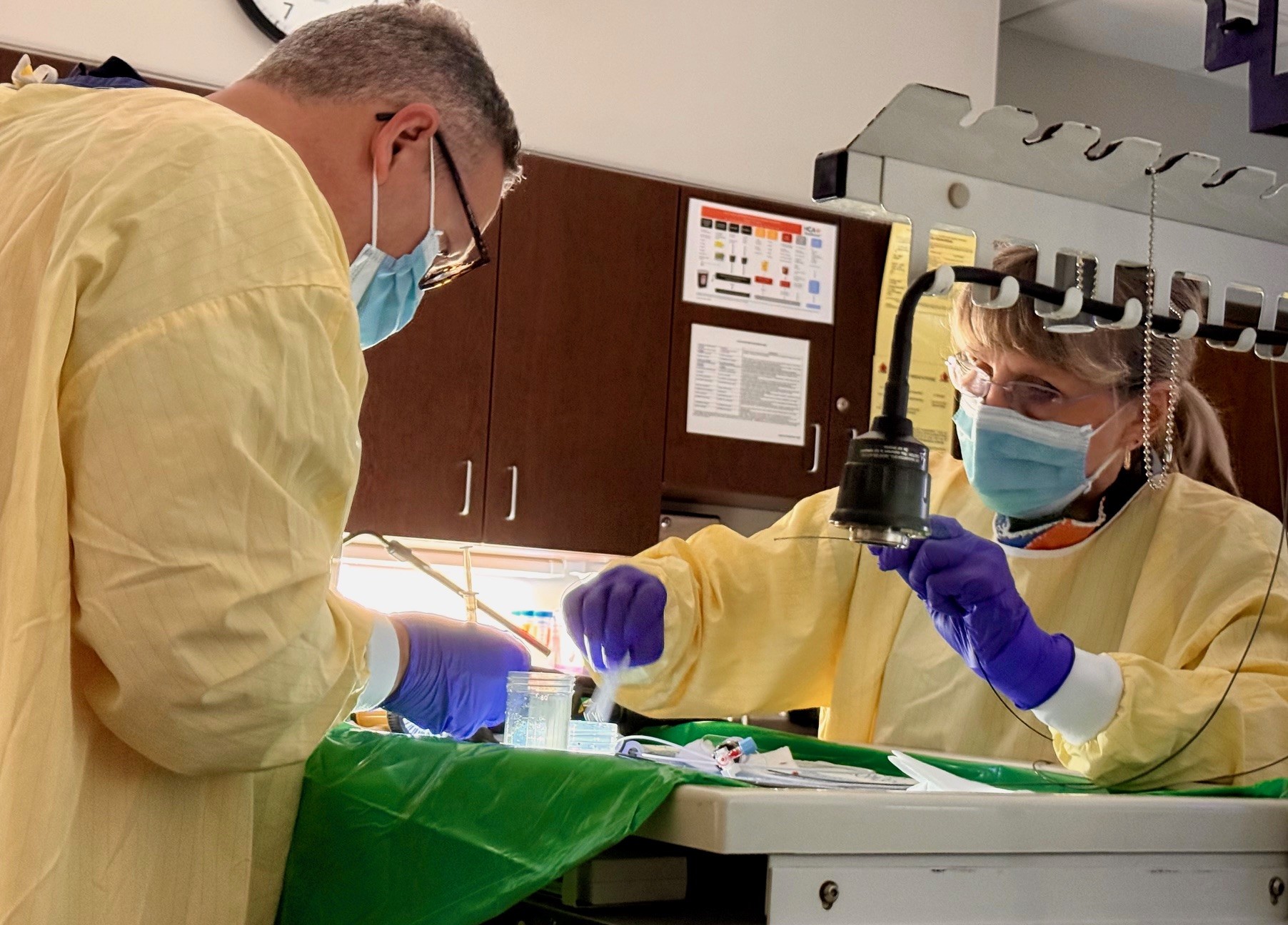 Two healthcare workers wearing masks, gloves, and protective gowns prepare medical materials on a tray under a task light in a clinical setting.