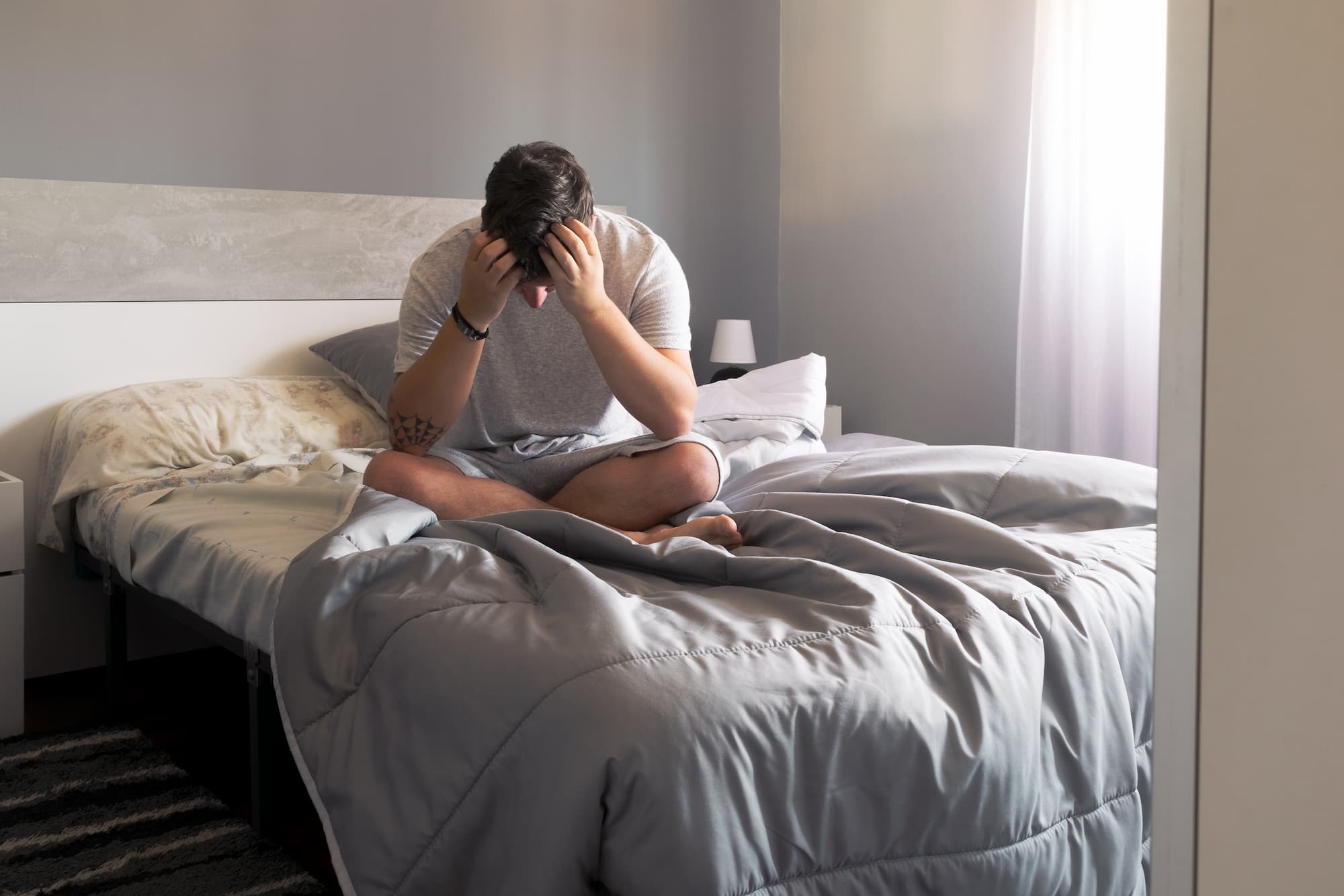 A man holds his head in his hands while sitting in bed.