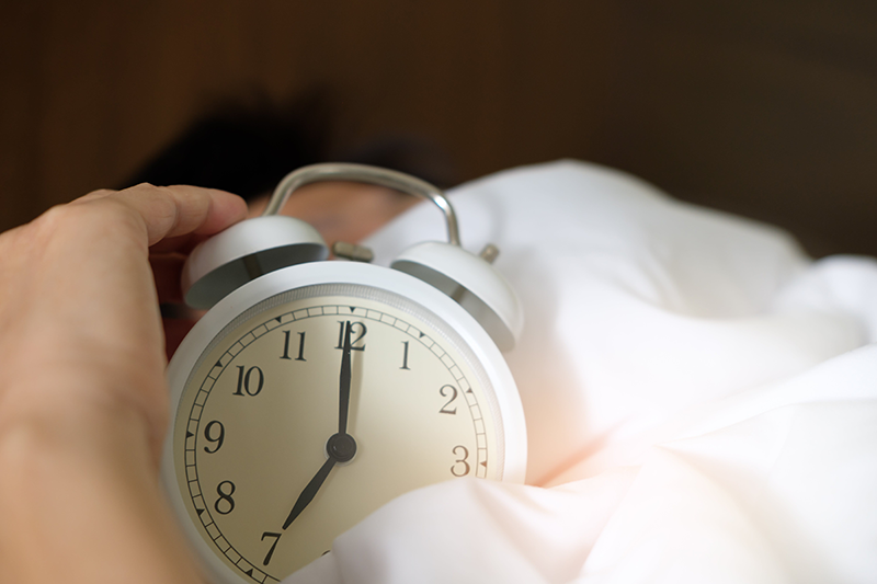 A person holds up a twin bell alarm clock while laying in bed.