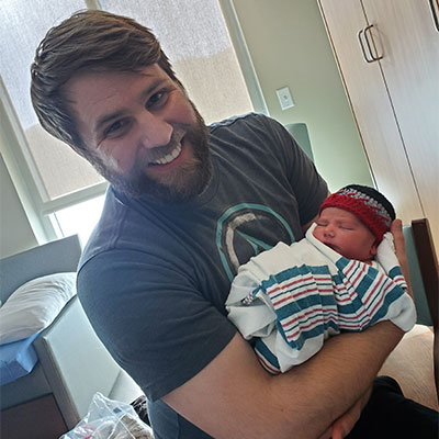 Josh Hathaway smiling as he holds his swaddled newborn son in a hospital room.