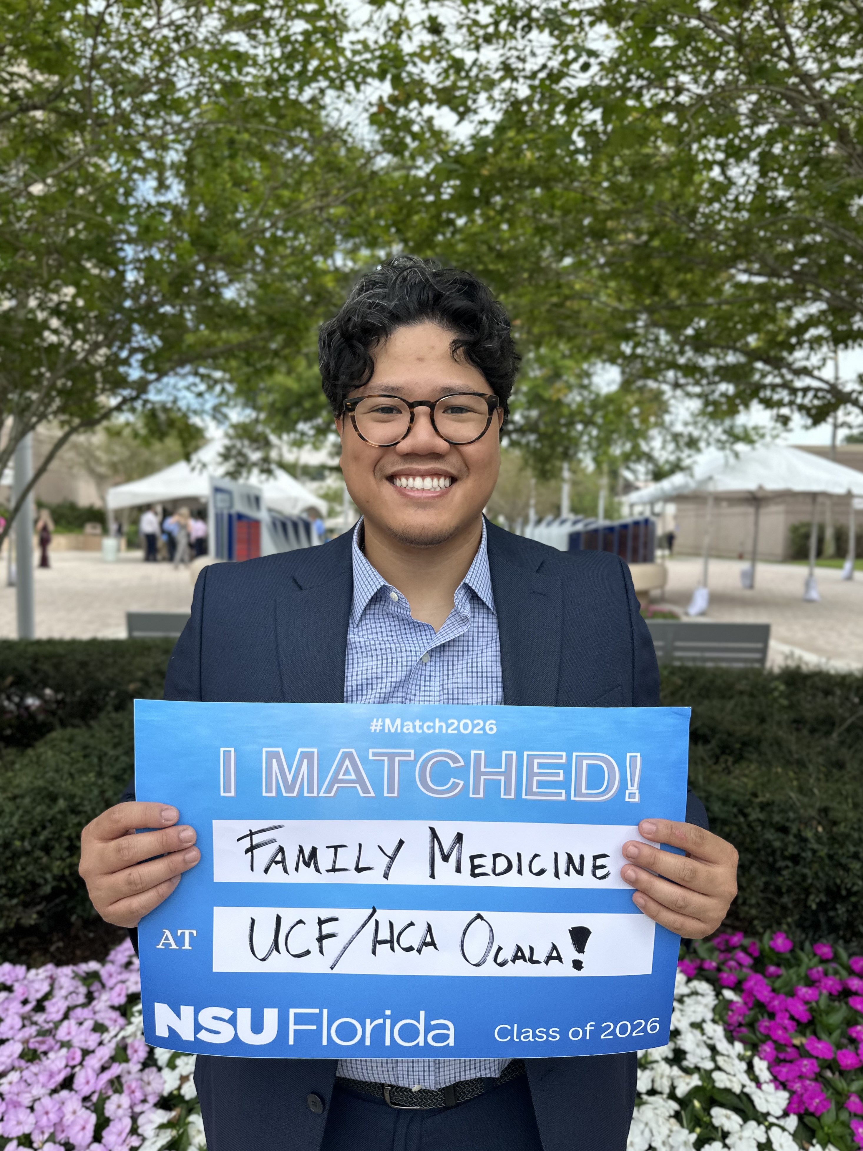 A smiling person wearing glasses and a navy suit stands outdoors under leafy trees, holding a blue sign that reads "#Match2026 I MATCHED! Family Medicine at UCF/HCA Ocala! NSU Florida Class of 2026." White tents and a few people are visible in the background, with flowers in the foreground.
