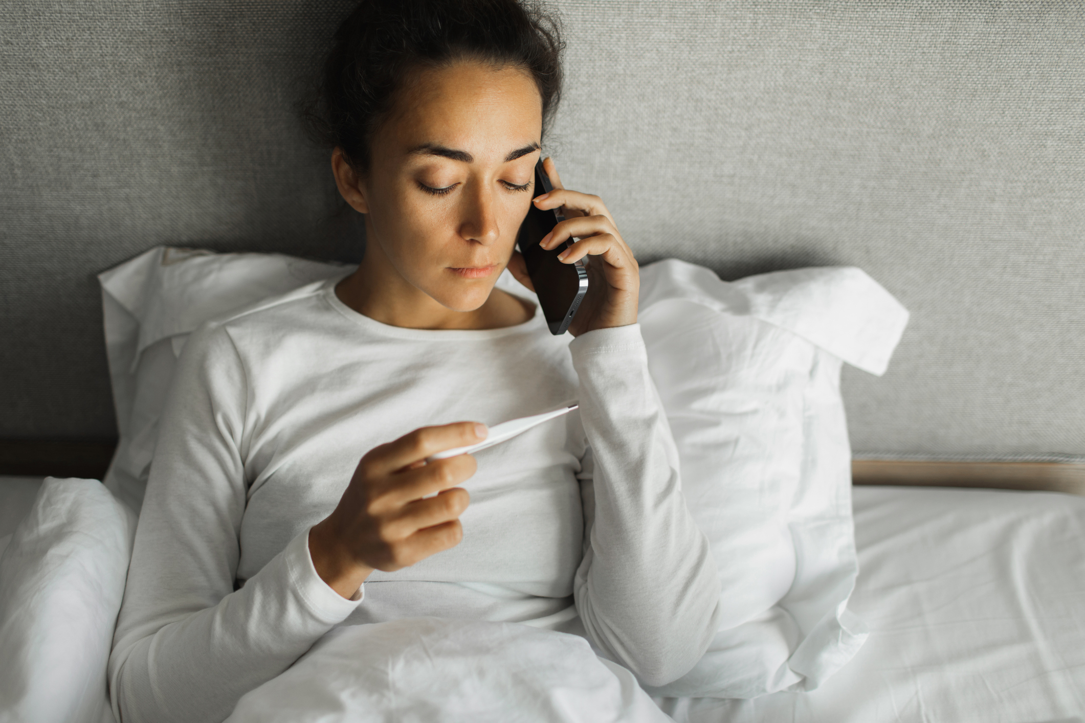 Worried woman sitting in bed with fever calling doctor to help her.