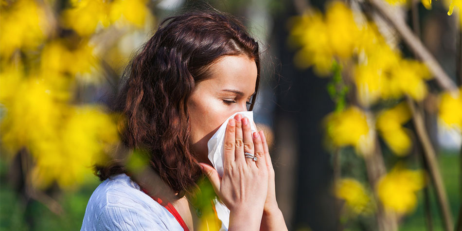 Woman blowing her nose outdoors surrounded by yellow flowers.