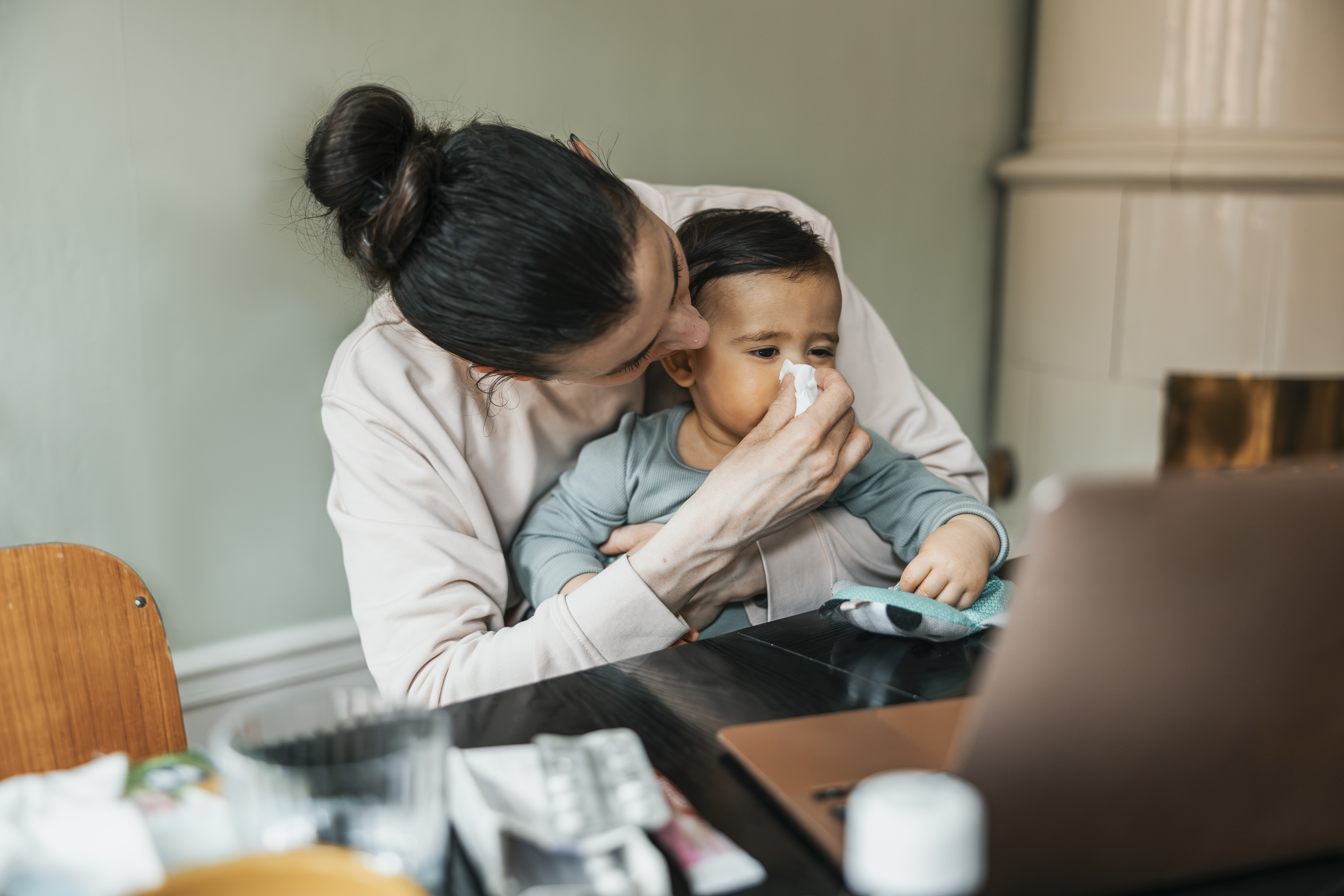 A mother holds her baby on her lap and wipes their nose with a tissue.