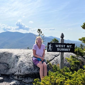 Carolyn sitting on a large rock, next to a sign that reads 'West Mt. Summit' on a hiking trail.