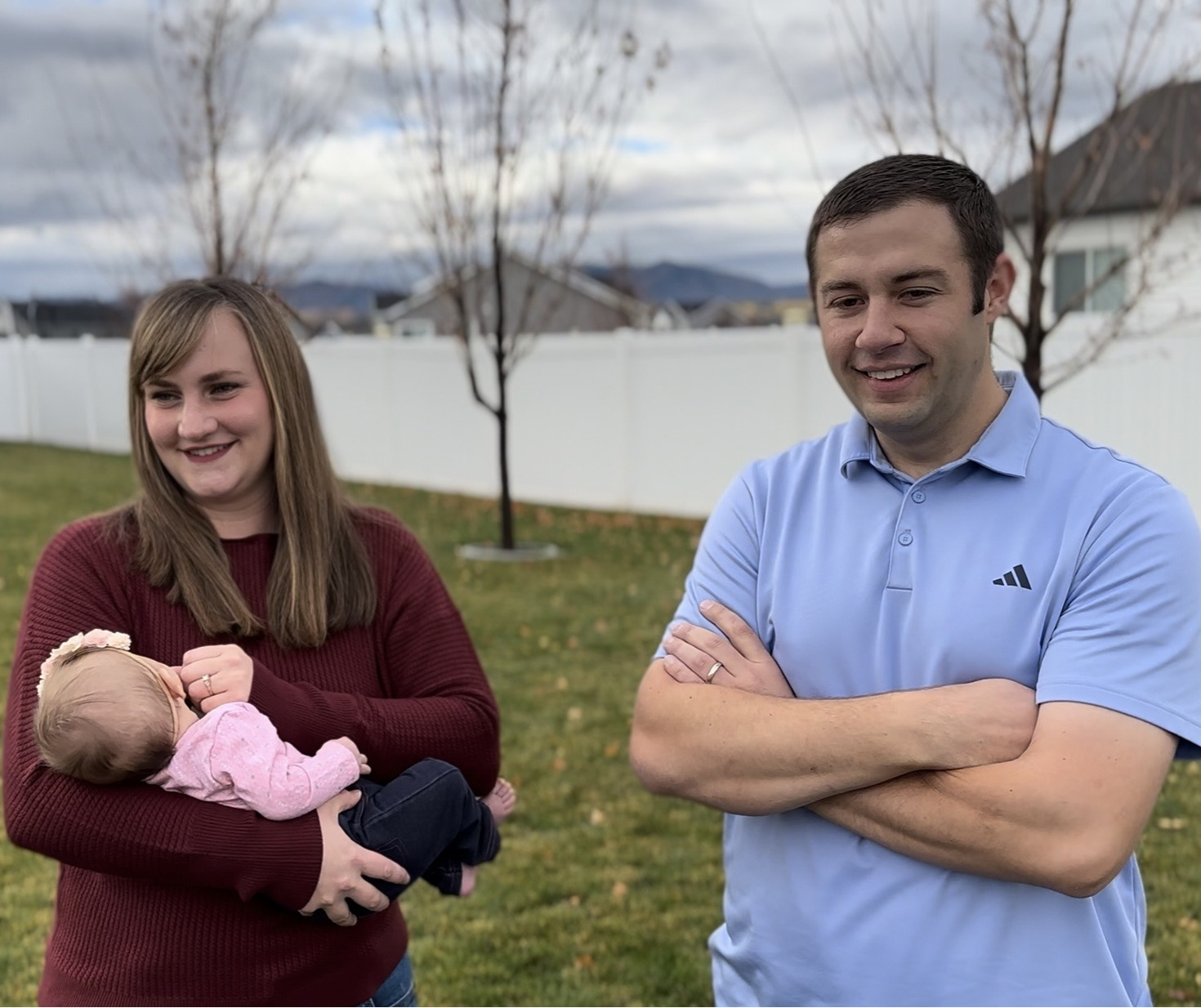 A woman holds a baby while standing next to a man in a backyard. Both are smiling, with houses, trees, and mountains visible in the background.