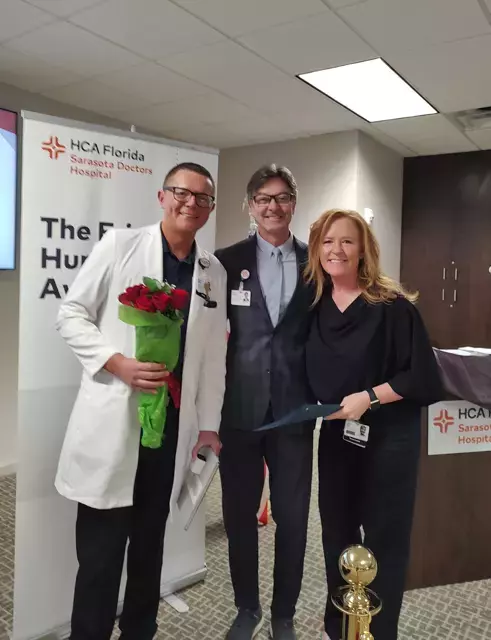 Woman stands smiling with two men holding award and flowers