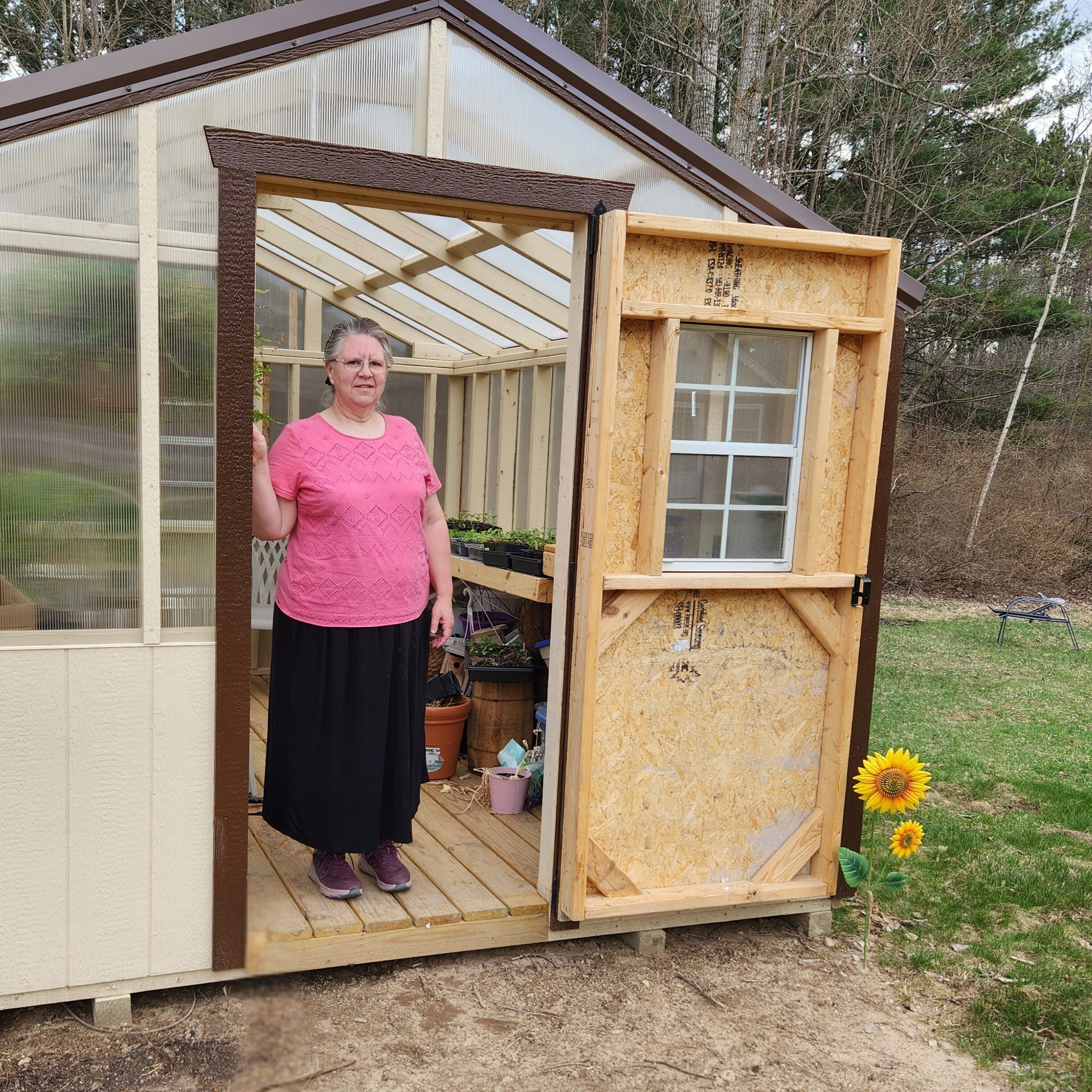 Hannah Martin stands in the doorway of a small greenhouse, surrounded by potted plants and garden tools.