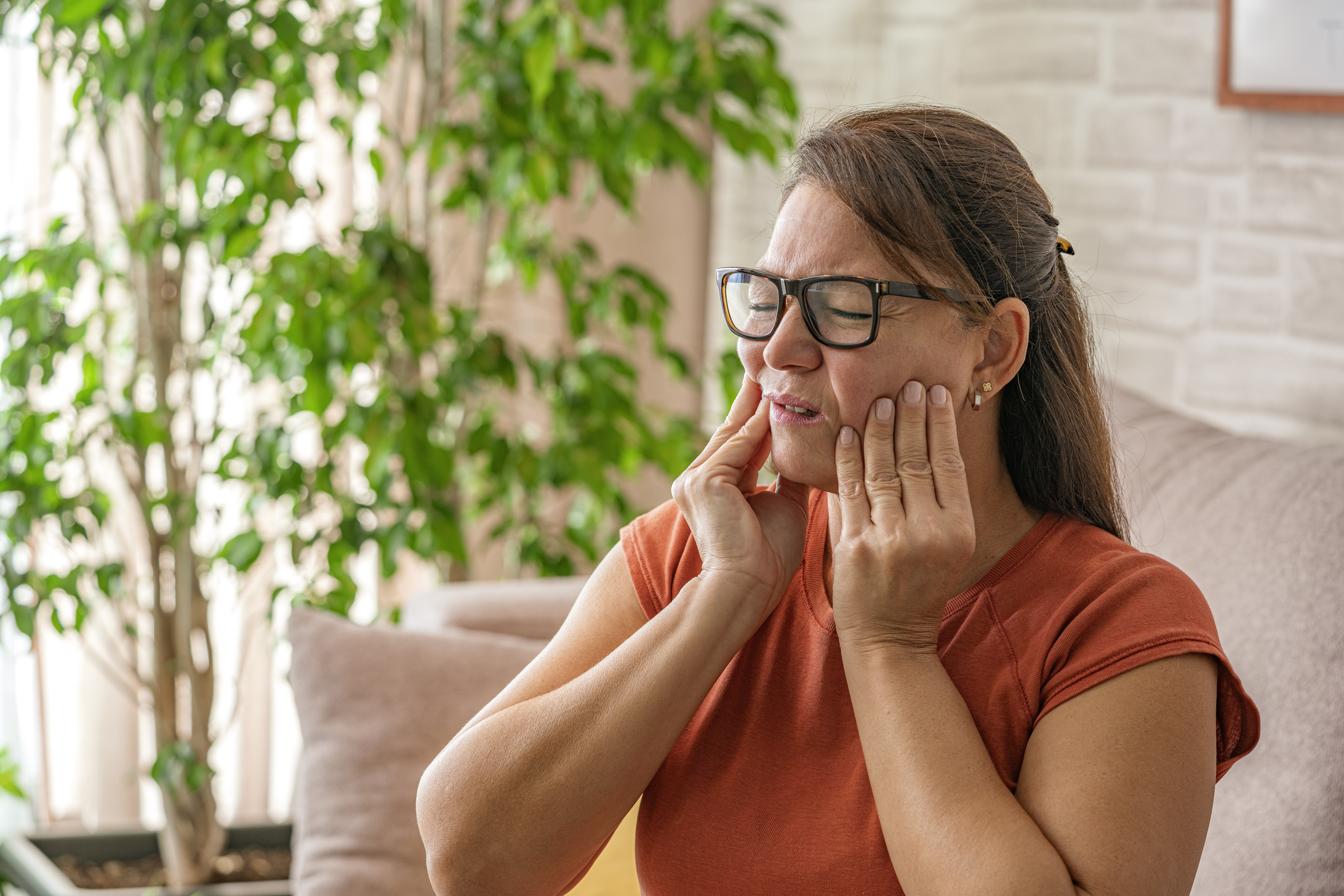 A woman sitting on a sofa with severe dental pain, holding her hands to her face.